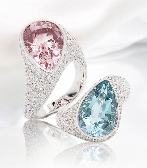 Two rings side by side. One has a large, teardrop - shaped pink gemstone as the centerpiece, surrounded by a halo of tiny, glittering diamonds. The other features a similarly sized teardrop - shaped blue gemstone, also encircled by a cluster of shining diamonds.