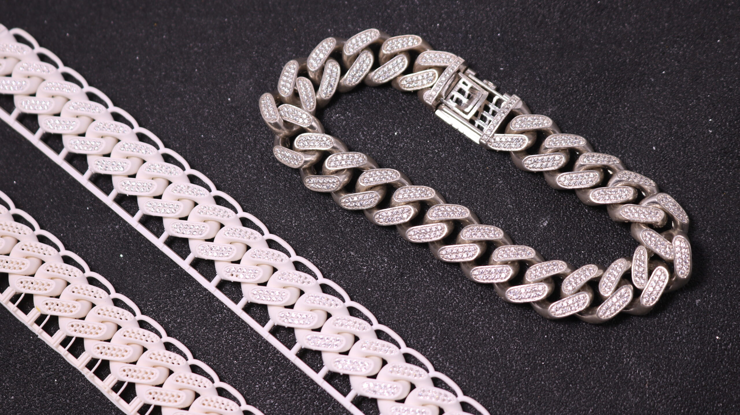 On a dark textured surface, there's a shiny silver - cast bracelet with large, diamond - encrusted oval links and a detailed clasp. Next to it lie two white, 3D - printed bracelets made of white wax resin. These 3D - printed pieces have a similar linked structure to the silver one, with small, simulated diamond - like elements embedded in each link, and a more delicate, lightweight appearance.