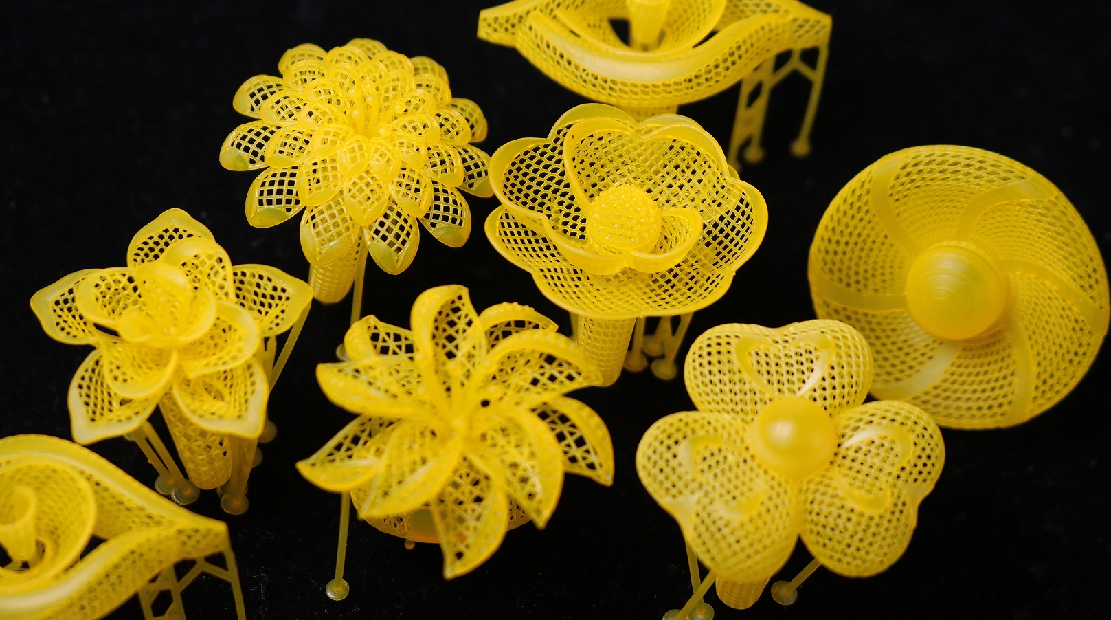 A collection of 3D - printed jewelry pieces in a vivid yellow color, inspired by flowers. Each piece has a delicate, mesh - like structure, with some resembling blooming petals and others having a circular, fan - like shape. They stand on thin supports against a dark black background, giving an impression of floating, intricate floral artworks.