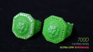 Two vibrant green 3D - printed rings sit against a dark backdrop. Each ring has an octagonal face with a detailed lion head carving in the center. The left ring is bordered by a row of shiny, diamond - like embellishments, while the right ring features a border of small holes and raised dots, adding texture to its unique design. The text "700D CASTING RESIN ULTRA LOW SHRINKAGE" is visible near the bottom right.