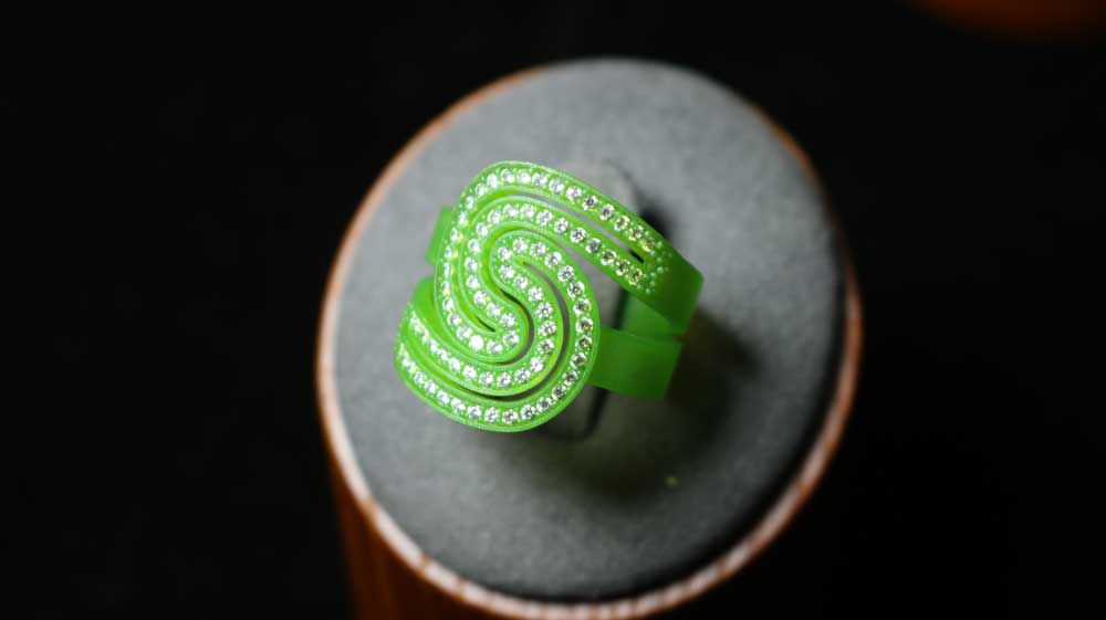 3D - printed green resin ring with a double - layered, spiral shaped band lined with small diamonds, displayed on a gray cushion