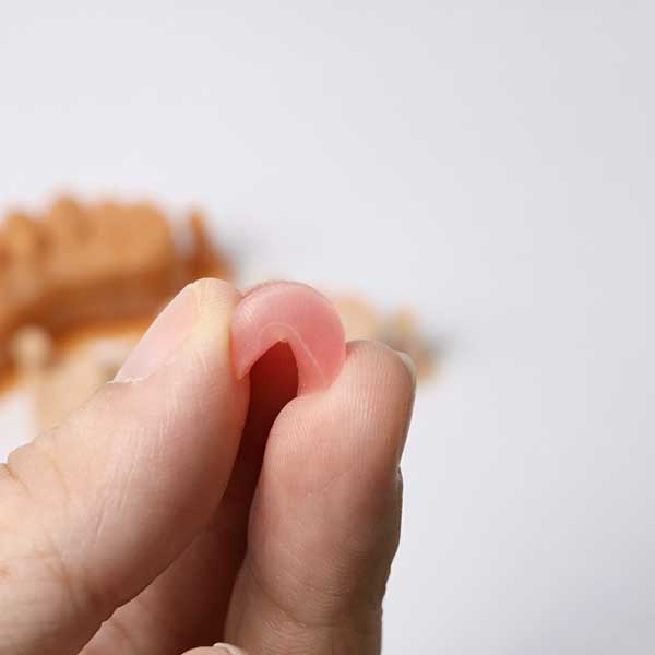 Fingers gently bending a pink gingiva mask, illustrating its silicone-like flexibility and excellent tear resistance.
