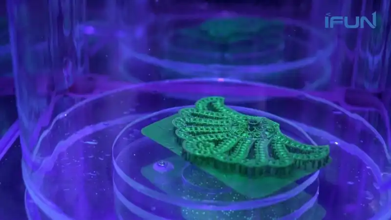 A detailed green resin 3D printed jewelry model with supports, placed on a transparent platform inside a UV curing chamber under purple light