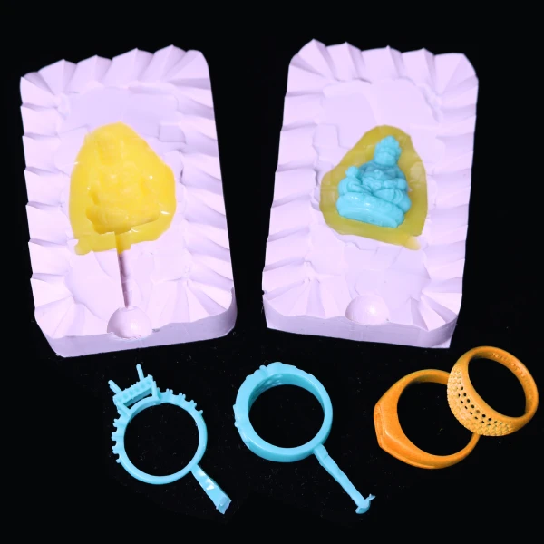 An open pink vulcanized rubber jewelry mold containing a blue wax Buddha, displayed with 3D printed orange master rings and intricate blue injected wax ring patterns with stone prongs.